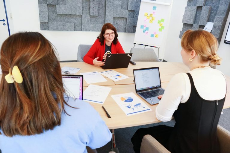 Professional business meeting scene in modern office, diverse team of professionals around conference table with laptops and documents, focused on AI training session, natural lighting through large windows, contemporary corporate atmosphere, business compliance concept, shot with professional camera, Pexels keywords: business meeting training corporate office — Formations 90